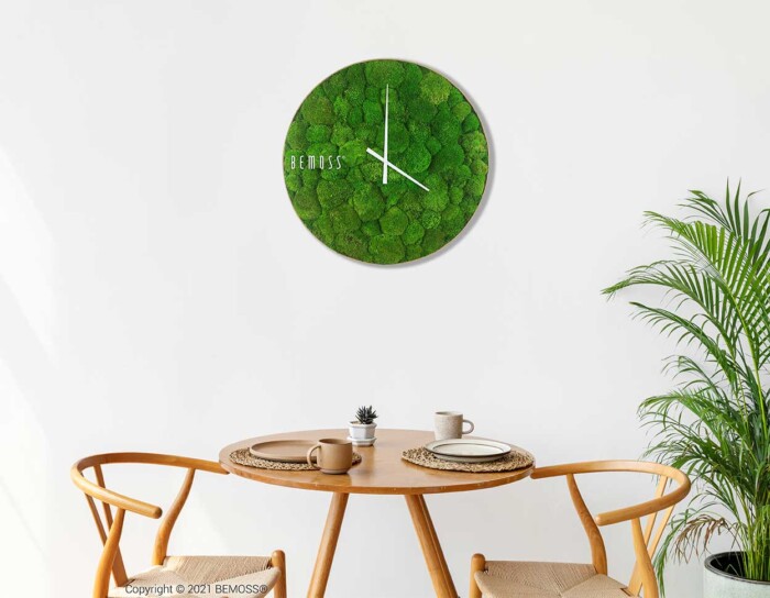 Eine grüne Wanduhr mit weißem Zifferblatt an einer weißen Wand über einem Tisch mit zwei Stühlen und einer Topfpflanze, moosbild, mooswand, moos pflanzen, moos, moos deko, moos art